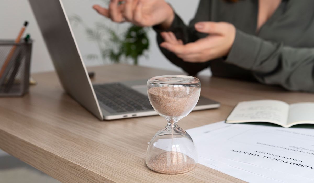 A woman sitting at a desk with a laptop and a sand timer, focused on her work in a well-lit office environment.