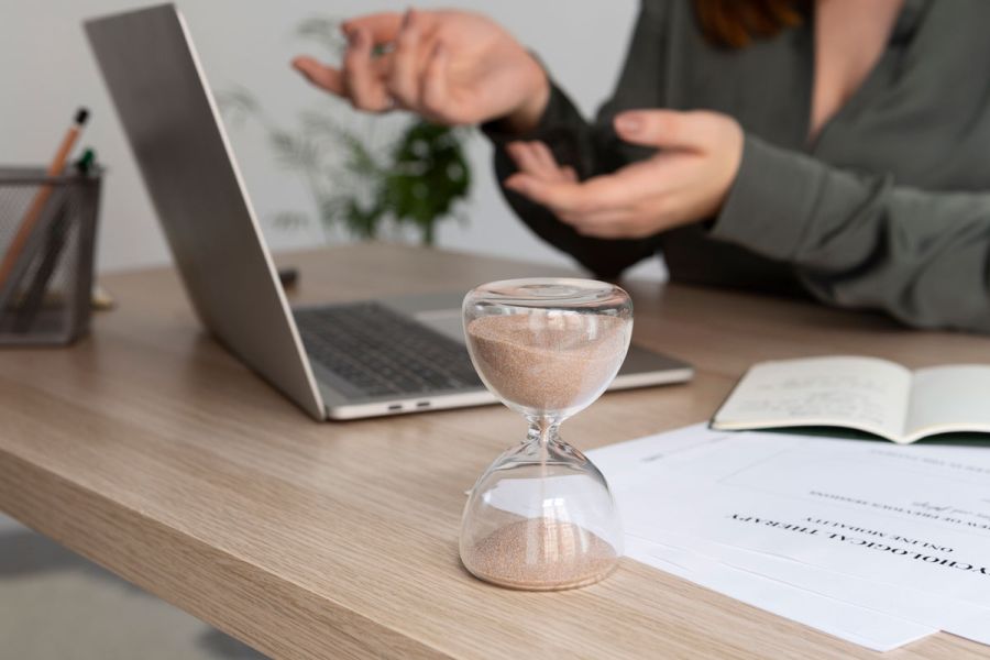 A woman sitting at a desk with a laptop and a sand timer, focused on her work in a well-lit office environment.
